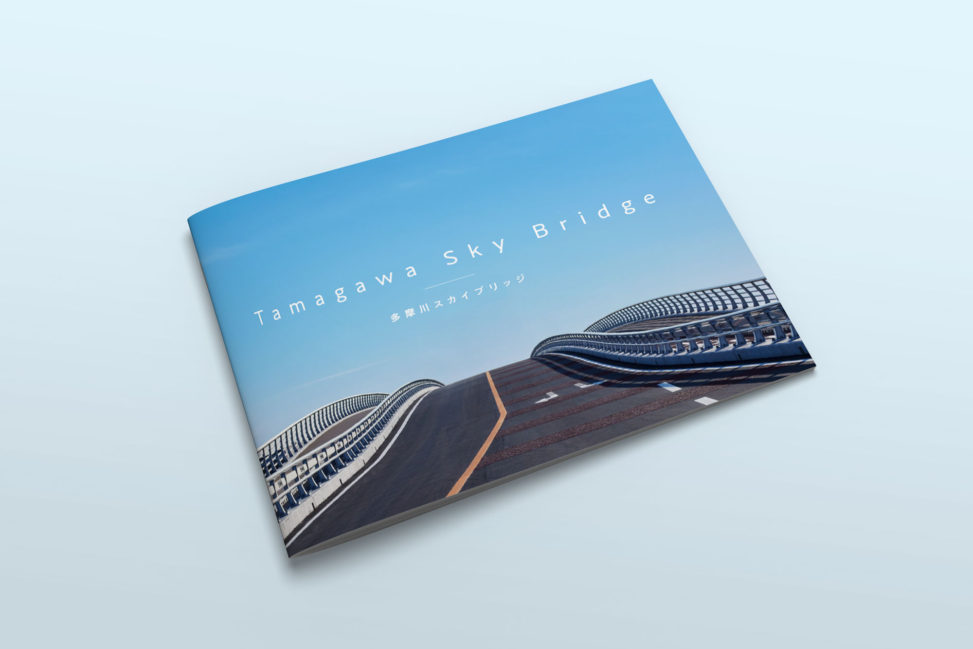 多摩川スカイブリッジの竣工パンフレット。青空の下、美しく設計された橋の曲線が際立つ表紙デザイン。パンフレットには、空撮や船上からの全景写真、ライトアップされた夕景などが掲載され、橋の特徴や魅力がわかりやすくまとめられている。開通式典で関係者に配布された公式資料。