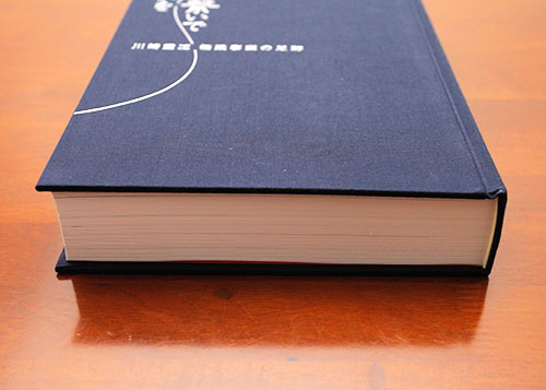 川崎重工の橋梁事業の足跡を記録した工事誌の側面写真。紺色のハードカバーに白いデザインが施され、高級感のある製本仕様。数百ページに及ぶ詳細な建設記録が収められ、企業の技術継承やアーカイブ用の書籍として作成された。