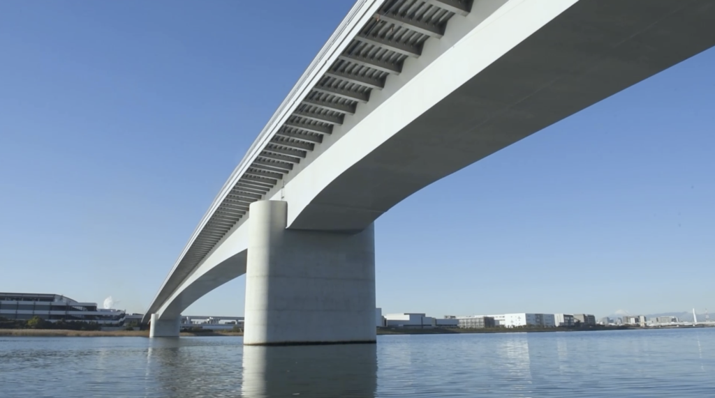 多摩川スカイブリッジの橋梁構造を水上から撮影。東京都大田区羽田と神奈川県川崎市殿町を結ぶ新たな公共工事。2022年春の開通を記念した映像制作の一環として、水面に映る橋の美しいシルエットと周囲の都市景観を記録。青空の下での壮大な橋の全景を紹介。